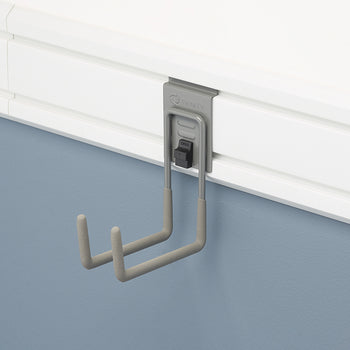 A close-up shot of a gray, U-shaped hook hanging from a white slatted wall panel. The hook, which has two prongs, is designed to hold various items and is attached to the panel via a small, black locking mechanism.