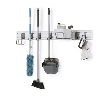 A modular wall-mounted storage system holds various cleaning tools on a white wall. The gray rack organizes a blue duster, a black broom, a gray dustpan and broom set, and a caddy with cleaning supplies, brushes, and a towel.