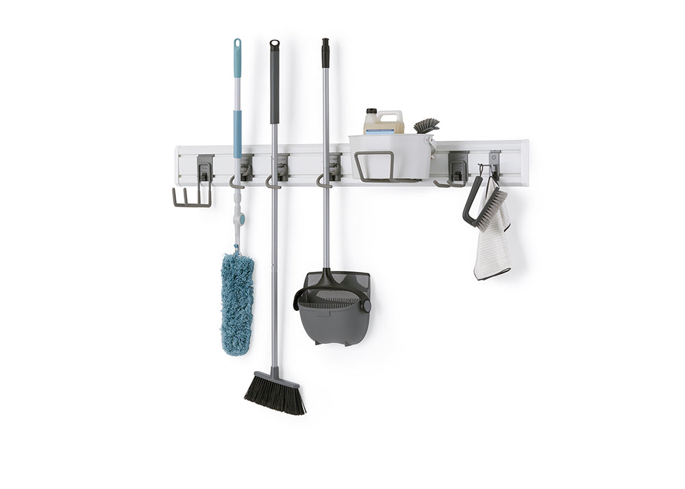 A modular wall-mounted storage system holds various cleaning tools on a white wall. The gray rack organizes a blue duster, a black broom, a gray dustpan and broom set, and a caddy with cleaning supplies, brushes, and a towel.