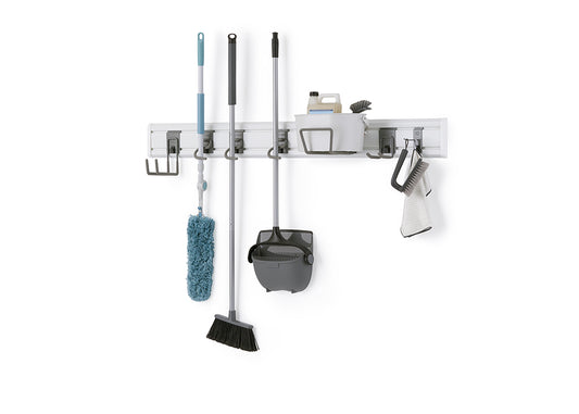 A modular wall-mounted storage system holds various cleaning tools on a white wall. The gray rack organizes a blue duster, a black broom, a gray dustpan and broom set, and a caddy with cleaning supplies, brushes, and a towel.