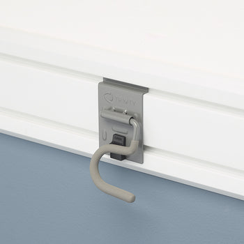 A close-up shot of a gray TRINITY brand hook mounted on a white slatwall panel. The hook has a curved, rubberized end and a spring-loaded latch to secure it in place.