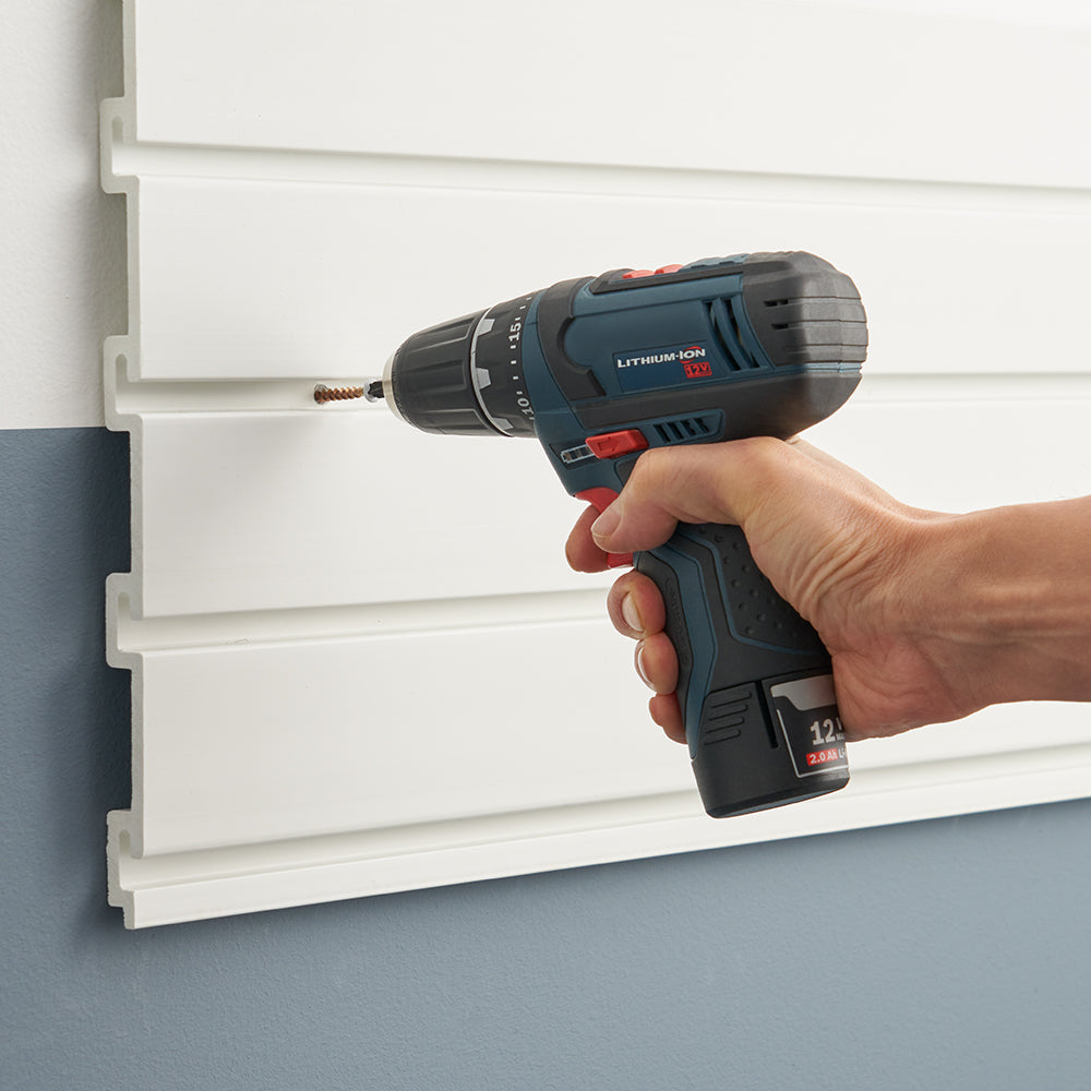A person is using a cordless power drill to drive a screw into a white slatwall panel mounted on a wall. The drill is blue and black with a lithium-ion battery, and the person's hand is gripping the handle firmly.