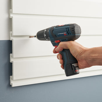 A person is using a cordless power drill to drive a screw into a white slatwall panel mounted on a wall. The drill is blue and black with a lithium-ion battery, and the person's hand is gripping the handle firmly.