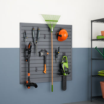A garage or shed wall features a dark gray slatwall system holding various gardening tools. To the right, a metal shelving unit holds terracotta pots, bags of soil, and a green watering can.