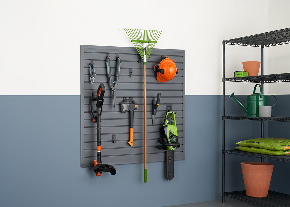 A garage or shed wall features a dark gray slatwall system holding various gardening tools. To the right, a metal shelving unit holds terracotta pots, bags of soil, and a green watering can.