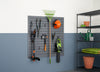 A garage or shed wall features a dark gray slatwall system holding various gardening tools. To the right, a metal shelving unit holds terracotta pots, bags of soil, and a green watering can.