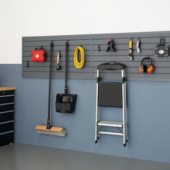 A well-organized garage features a gray slatwall system holding various tools and equipment, including a broom, shovel, garden hose, and safety gear. To the left, a black storage cabinet with a wooden top provides additional workspace and storage.