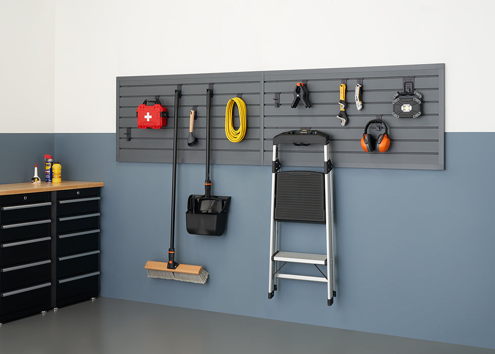 A well-organized garage features a gray slatwall system holding various tools and equipment, including a broom, shovel, garden hose, and safety gear. To the left, a black storage cabinet with a wooden top provides additional workspace and storage.