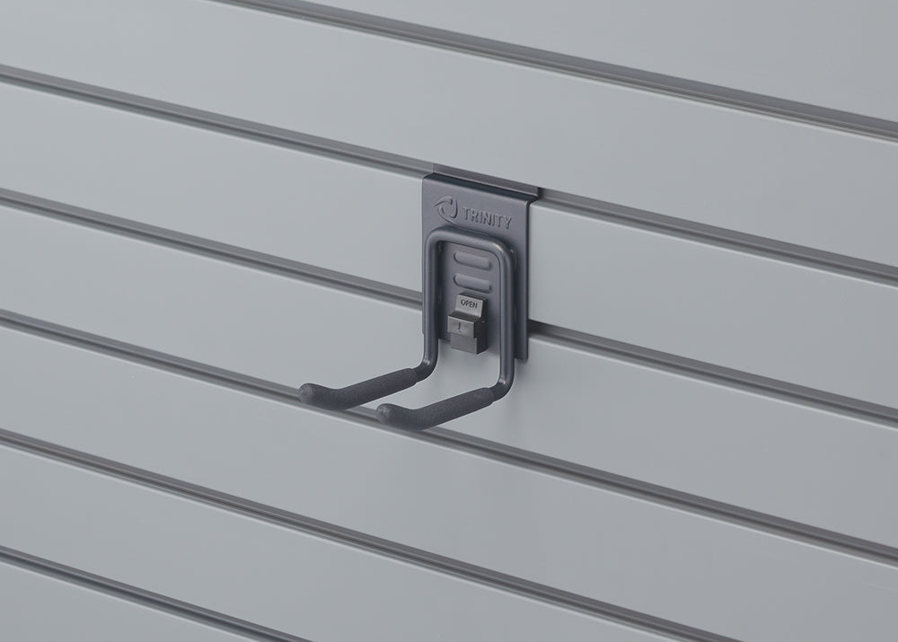 A close-up shot shows a black, U-shaped hook with "TRINITY" embossed on its top, attached to a light gray slat wall. The hook has two prongs extending forward and a small, gray "OPEN" tab visible on its base.