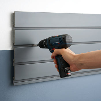 A person uses a blue and black cordless drill to screw into a gray slat wall panel. The panel is mounted on a two-toned wall, with the upper section being gray and the lower section blue.