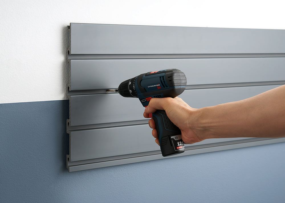 A person uses a blue and black cordless drill to screw into a gray slat wall panel. The panel is mounted on a two-toned wall, with the upper section being gray and the lower section blue.