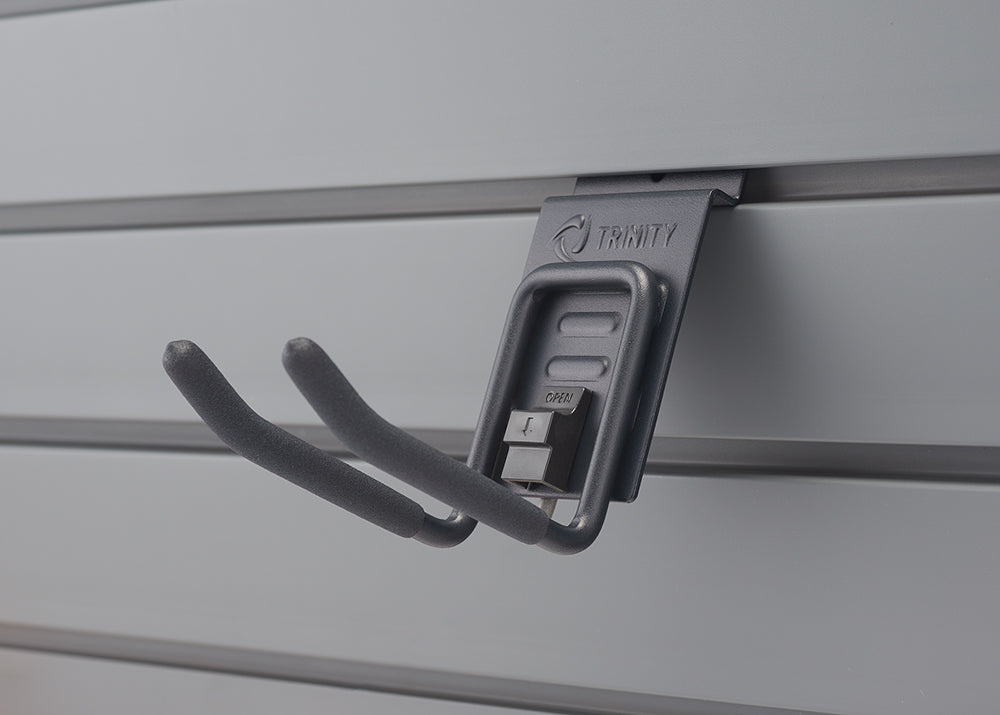A close-up shot shows a gray double-prong hook with foam padding on the prongs, hanging from a gray slatwall panel. The hook has "TRINITY" embossed at the top and a small "OPEN" lever for adjustment.