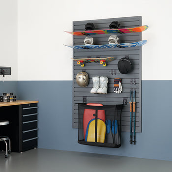 A neatly organized garage features a gray slatted wall storage system holding two snowboards, a skateboard, winter sports gear like a helmet, boots, and gloves, and a mesh bag with what appears to be water sports equipment. To the left, a black workbench with drawers and a silver stool are visible, all against a wall painted white on top and a muted blue-gray on the bottom.