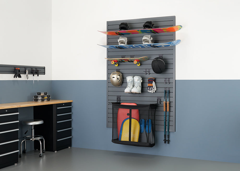 A neatly organized garage features a gray slatted wall storage system holding two snowboards, a skateboard, winter sports gear like a helmet, boots, and gloves, and a mesh bag with what appears to be water sports equipment. To the left, a black workbench with drawers and a silver stool are visible, all against a wall painted white on top and a muted blue-gray on the bottom.