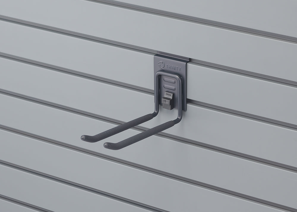 A close-up shot shows a gray double-prong hook with a black "TRINITY" logo attached to a light gray slatwall. The hook has an "OPEN" latch at the top, allowing it to be securely fastened or removed from the slatwall.