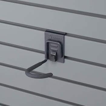 A Trinity brand utility hook is mounted on a gray slat wall. The hook is dark gray with a black foam-covered grip, and a small black latch on its base is labeled 