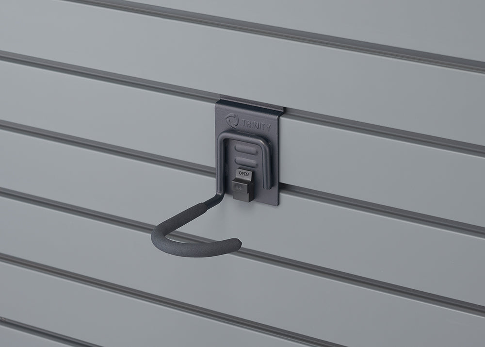 A Trinity brand utility hook is mounted on a gray slat wall. The hook is dark gray with a black foam-covered grip, and a small black latch on its base is labeled "OPEN."
