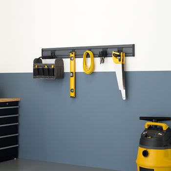 A well-organized garage or workshop with a gray and white painted wall. A tool rack mounted on the wall holds a saw, a level, and a tool bag, while a yellow and black shop vacuum and a yellow work light are on the floor.