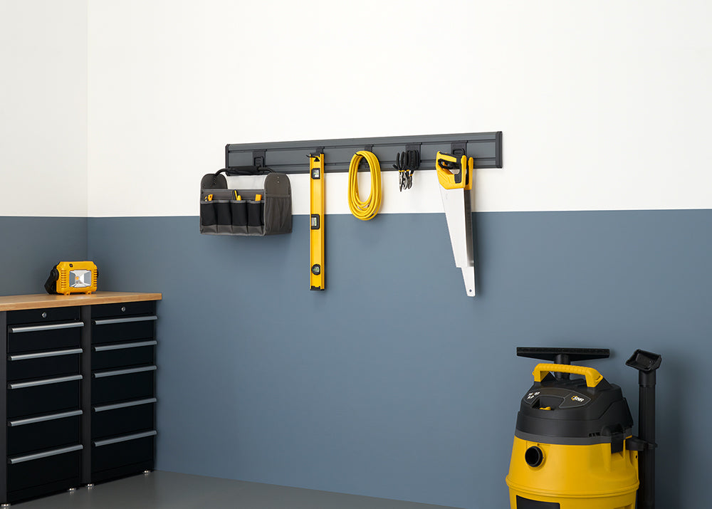 A well-organized garage or workshop with a gray and white painted wall. A tool rack mounted on the wall holds a saw, a level, and a tool bag, while a yellow and black shop vacuum and a yellow work light are on the floor.