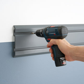 A person is using a Bosch 12V cordless drill to screw a metal track system into a wall. The track system, which appears to be a garage organization solution, is being installed on a wall that is painted white on top and blue on the bottom.