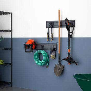 A neatly organized storage wall for gardening and yard tools. Tools like a shovel, a small weed trimmer, a watering can, a hose, and a pair of pliers are hanging from a dark gray rack mounted on a two-tone wall.