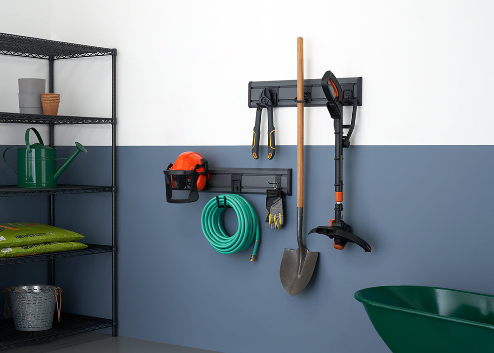 A neatly organized storage wall for gardening and yard tools. Tools like a shovel, a small weed trimmer, a watering can, a hose, and a pair of pliers are hanging from a dark gray rack mounted on a two-tone wall.
