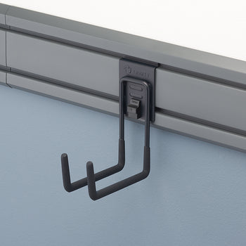 A close-up shot of a gray storage hook, with two J-shaped arms, attached to a rail system mounted on a light blue wall. The hook is part of the Trinity Pro Series and has a mechanism with 