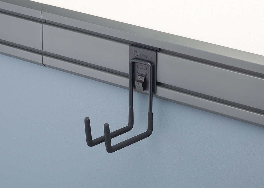 A close-up shot of a gray storage hook, with two J-shaped arms, attached to a rail system mounted on a light blue wall. The hook is part of the Trinity Pro Series and has a mechanism with "OPEN" text visible, suggesting it can be easily attached or removed from the rail.