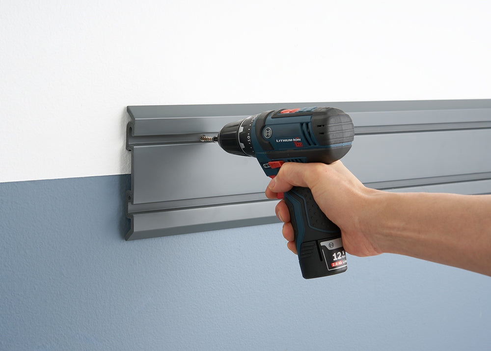A hand holds a blue and black cordless drill, driving a screw into a gray, slotted wall panel. The wall is painted white on top and a dark blue-gray on the bottom.