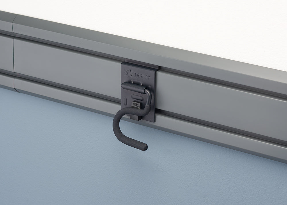 A close-up shot of a gray storage hook, labeled "TRINITY," attached to a slotted mounting rail on a wall. The hook has a curved end and is shown in the open position, with the word "OPEN" visible on the hook's base.