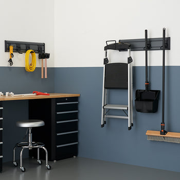 A garage workspace is organized with a workbench, a stool, and various tools. A wall-mounted rack holds a ladder, a dustpan, and a broom, while another smaller rack organizes a variety of hand tools and electrical cords.
