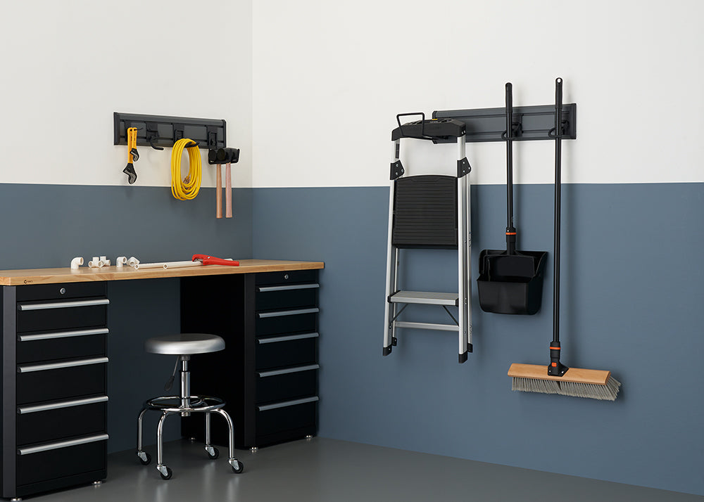 A garage workspace is organized with a workbench, a stool, and various tools. A wall-mounted rack holds a ladder, a dustpan, and a broom, while another smaller rack organizes a variety of hand tools and electrical cords.