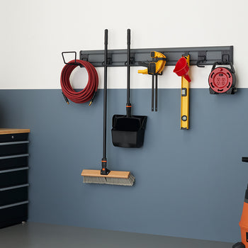 A well-organized garage wall featuring a storage system with various tools and equipment hanging neatly. A red hose, a broom and dustpan, a clamp, a level, and an extension cord are all suspended from a gray rail-like rack.