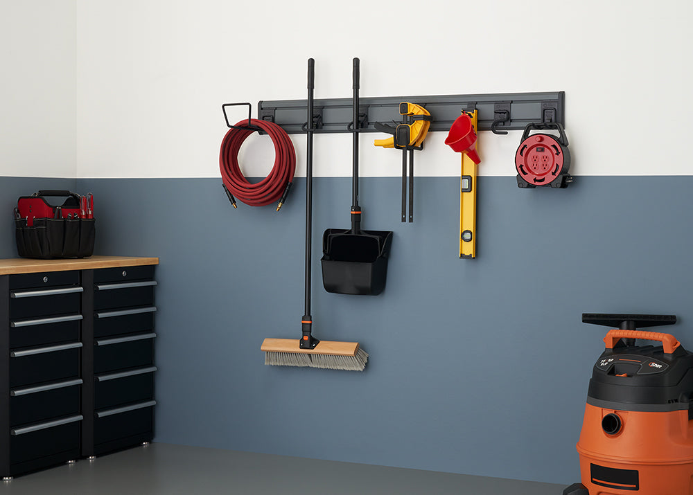 A well-organized garage wall featuring a storage system with various tools and equipment hanging neatly. A red hose, a broom and dustpan, a clamp, a level, and an extension cord are all suspended from a gray rail-like rack.
