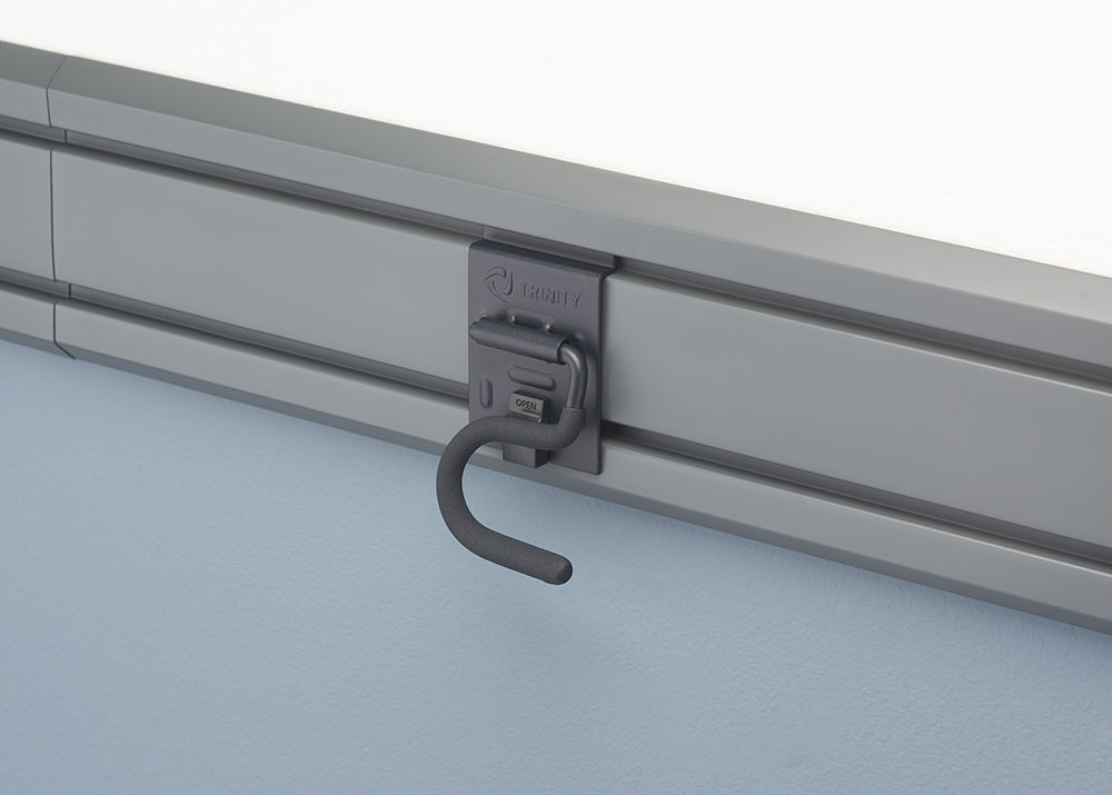 A close-up shot of a gray storage hook, branded "TRINITY," attached to a slotted wall track. The hook has a spring-loaded latch and is designed for hanging various items.