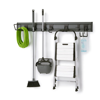 A dark gray, wall-mounted storage rack holds various cleaning tools and household items. From left to right, a green garden hose, a broom and dustpan set, a foldable step ladder, and a spray bottle are neatly hung on different hooks.