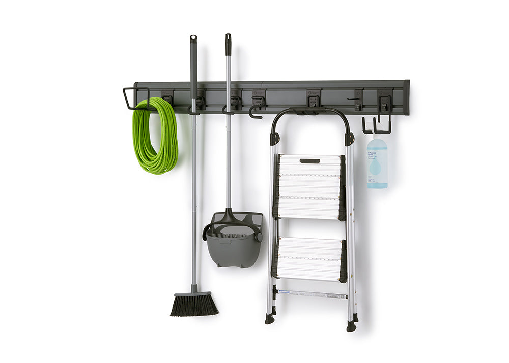 A dark gray, wall-mounted storage rack holds various cleaning tools and household items. From left to right, a green garden hose, a broom and dustpan set, a foldable step ladder, and a spray bottle are neatly hung on different hooks.