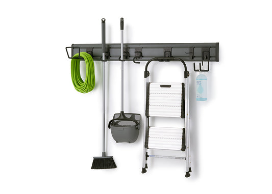 A dark gray, wall-mounted storage rack holds various cleaning tools and household items. From left to right, a green garden hose, a broom and dustpan set, a foldable step ladder, and a spray bottle are neatly hung on different hooks.