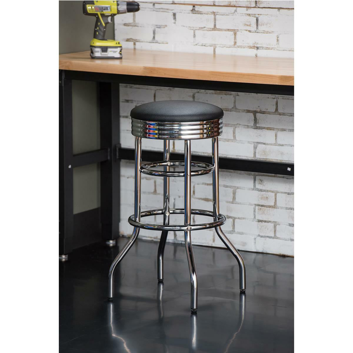 A chrome, 30-inch Trinity swivel bar stool with a black cushioned seat stands on a shiny black floor. In the background, there's a brick wall and a workbench with a drill.