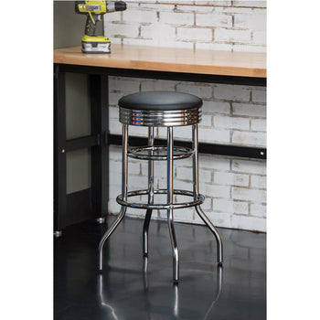 A chrome, 30-inch Trinity swivel bar stool with a black cushioned seat stands on a shiny black floor. In the background, there's a brick wall and a workbench with a drill.