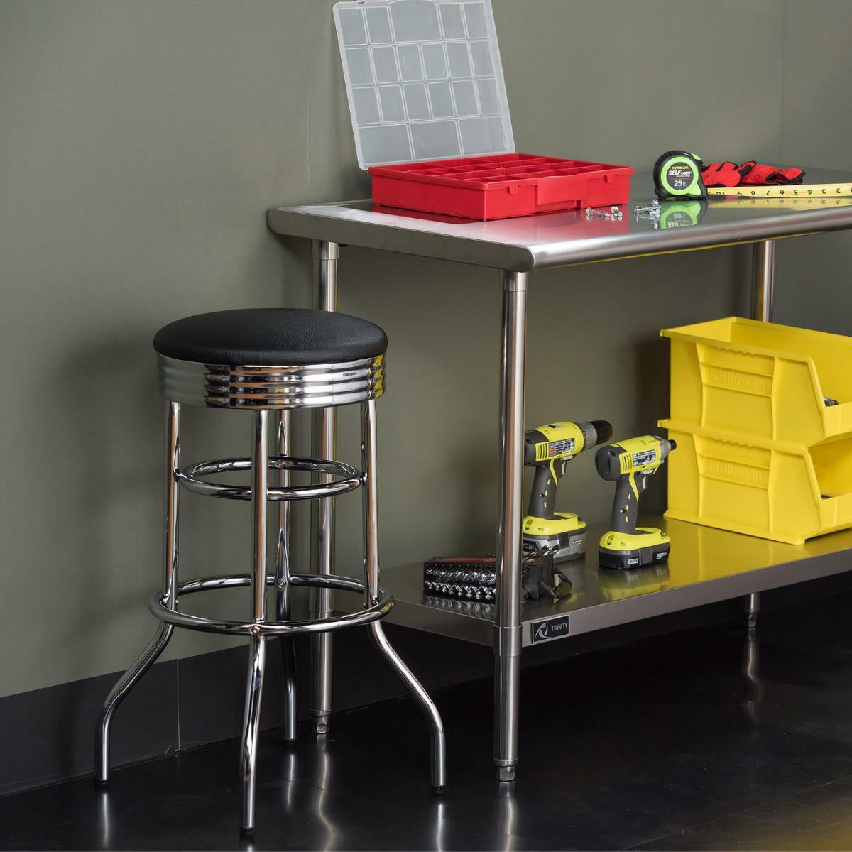 A chrome-legged stool with a black cushioned seat sits to the left of a stainless steel workbench. The workbench holds red and clear plastic organizers, a tape measure, and other tools, while a shelf below contains two yellow drills and other equipment.