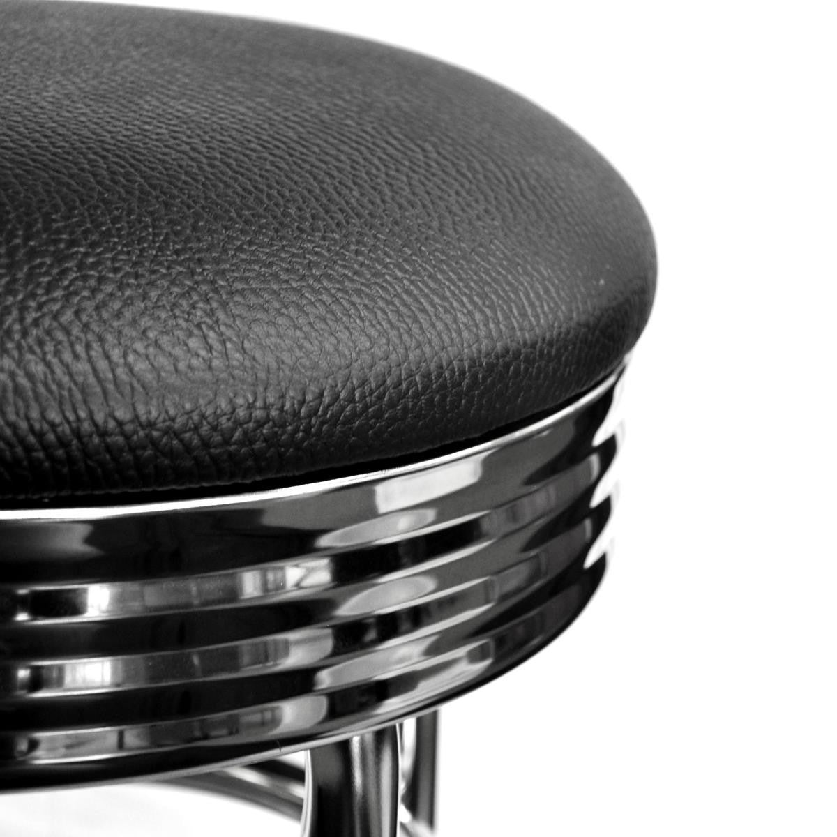 A close-up, black and white shot shows the upper portion of a vintage-style bar stool. The seat is upholstered in textured black material, and beneath it, a chrome-plated base features horizontal ribbed detailing.