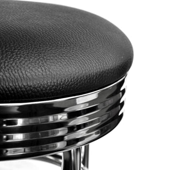 A close-up, black and white shot shows the upper portion of a vintage-style bar stool. The seat is upholstered in textured black material, and beneath it, a chrome-plated base features horizontal ribbed detailing.