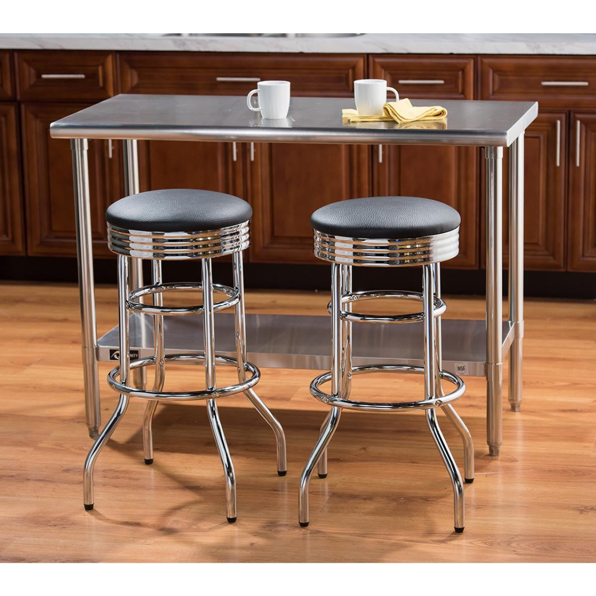 Two chrome swivel bar stools with black cushioned seats are positioned in front of a stainless steel kitchen island or table. The set includes a lower shelf on the table and is situated on a wooden floor against a backdrop of dark wood kitchen cabinets.