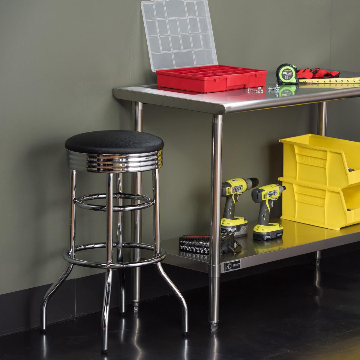 A workshop work stool beside a stainless steel work table. The work table has several work tools like two yellow drills, two yellow bins, a tape measure, and some small hardware on its top surface. 