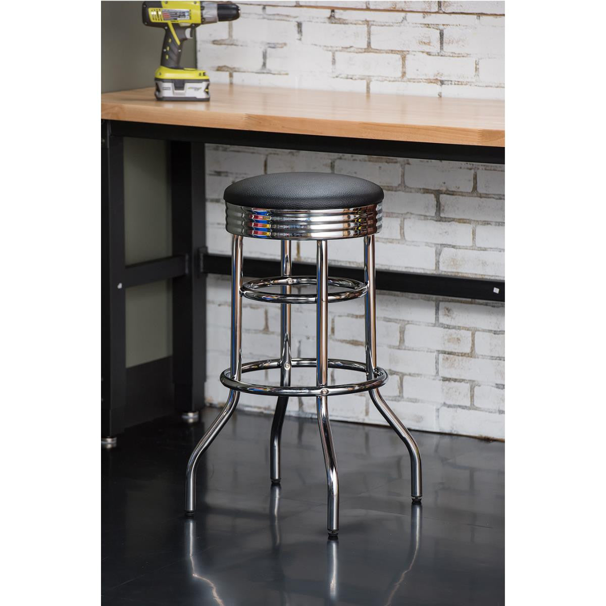 A chrome swivel bar stool with a black cushioned seat stands on a glossy black floor. Behind it is a light wood countertop against a white brick wall, where a yellow and black drill sits.
