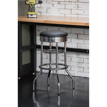 A chrome swivel bar stool with a black cushioned seat stands on a glossy black floor. Behind it is a light wood countertop against a white brick wall, where a yellow and black drill sits.