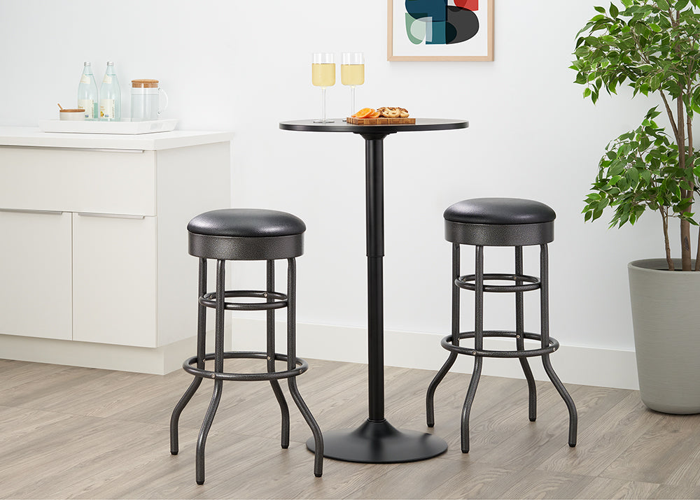 A small, black pub-style table with a round top stands between two matching tall bar stools. The stools have black faux leather seats and distressed black metal frames with curved legs.