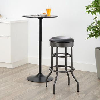 A dark, metal pub set is pictured, featuring a small, round pub table and a matching bar stool with a black, round cushion. The set is on a light wood floor in a brightly lit room with white cabinetry and a tall potted plant.