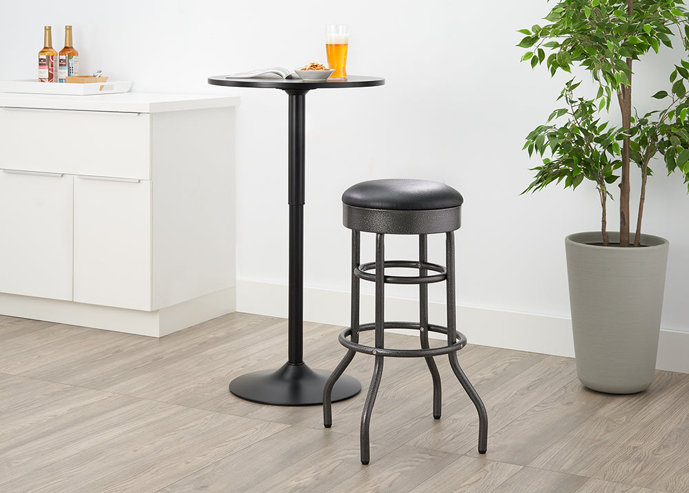 A dark, metal pub set is pictured, featuring a small, round pub table and a matching bar stool with a black, round cushion. The set is on a light wood floor in a brightly lit room with white cabinetry and a tall potted plant.